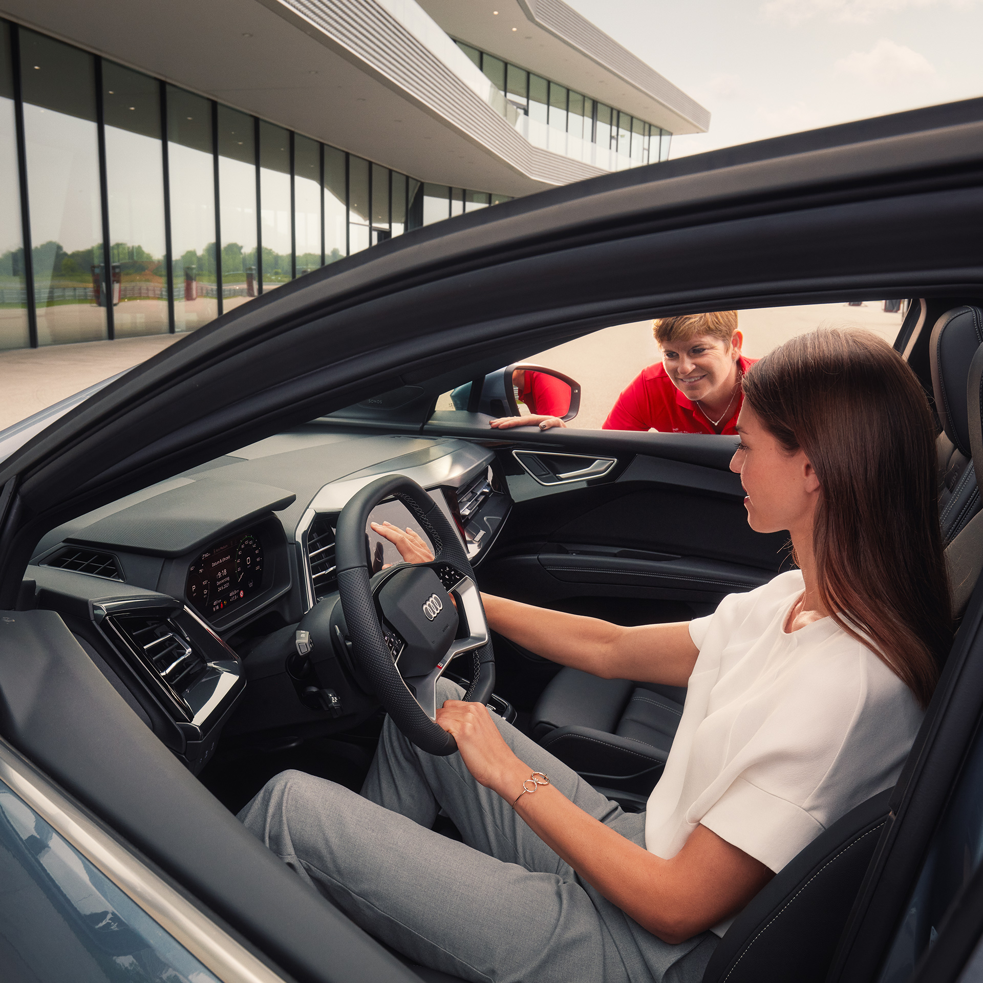 Frau mit dunklen langen Haarensitzt in einem Audi Modell, auf der Beifahrerseite schaut eine Frau in rotem T-Shirt durch das Fenster und erklärt das Fahrzeug