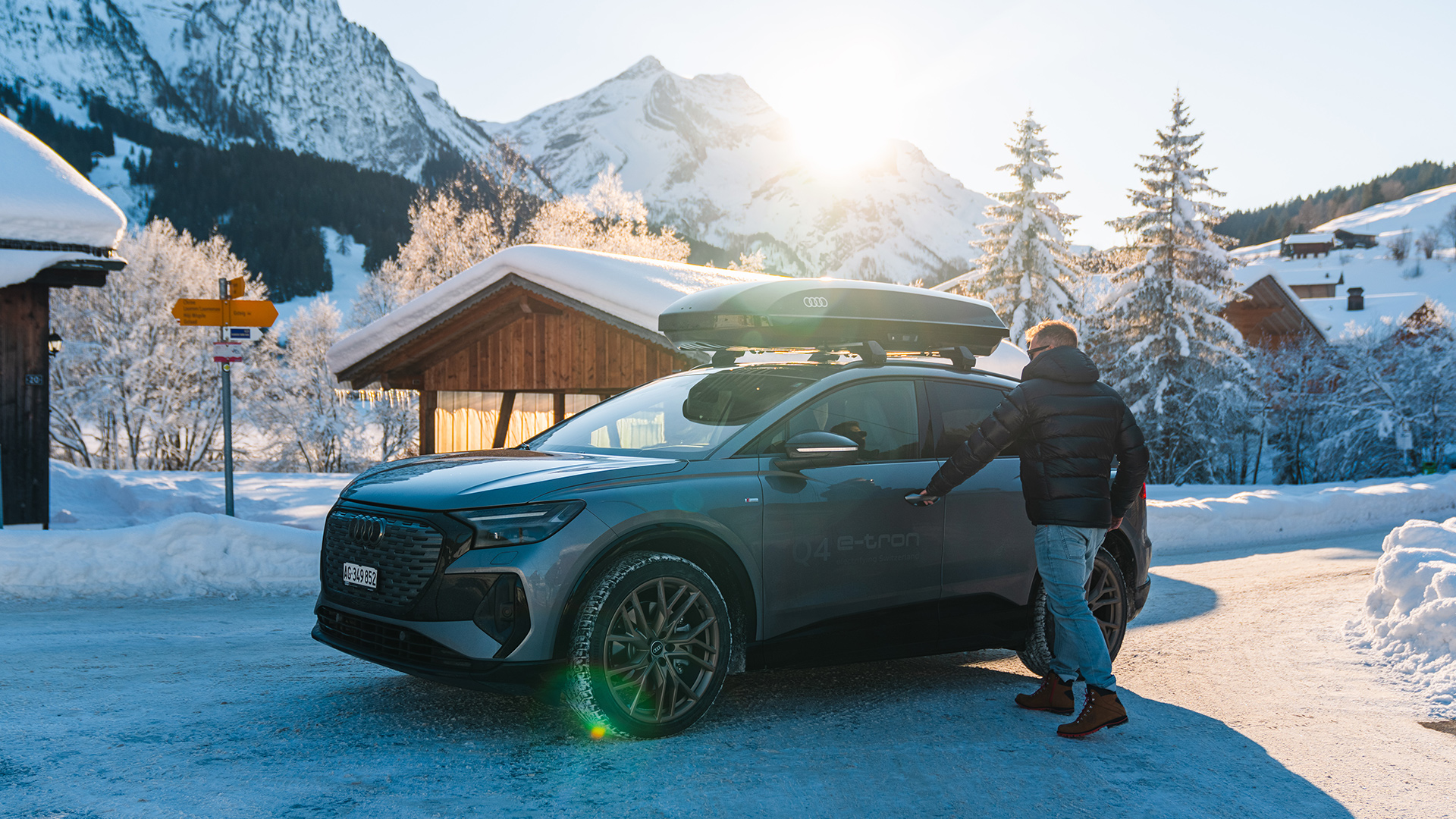 Silver Audi Q8 e-tron with luggage box stands in front of a wooden hut in the snow in the Alps, man wants to get into the car and open the driver's door