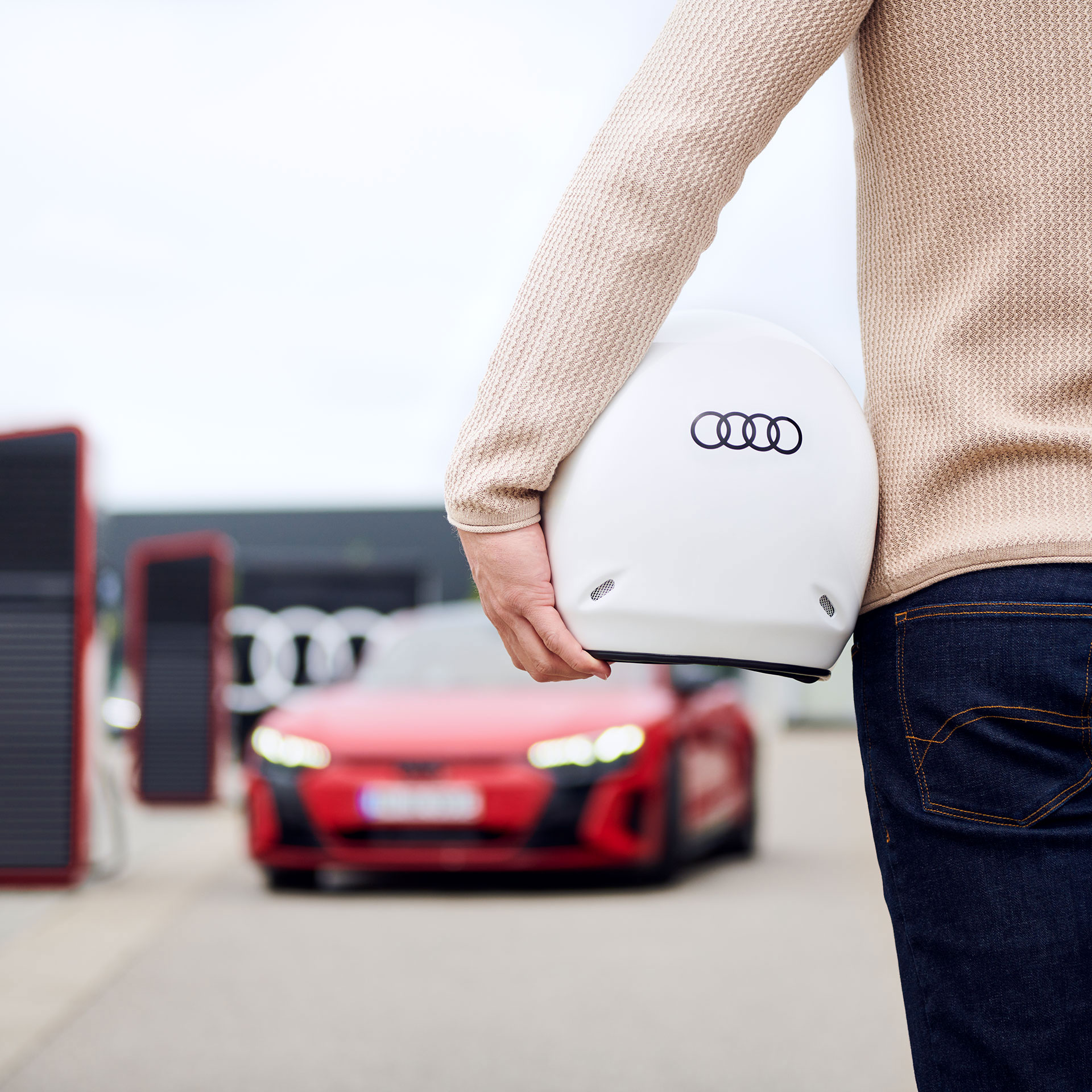 Rückansicht eines Mannes, der einen Helm mit Audi-Logo unter dem Arm trägt und auf ein rotes Audi Modell zugeht