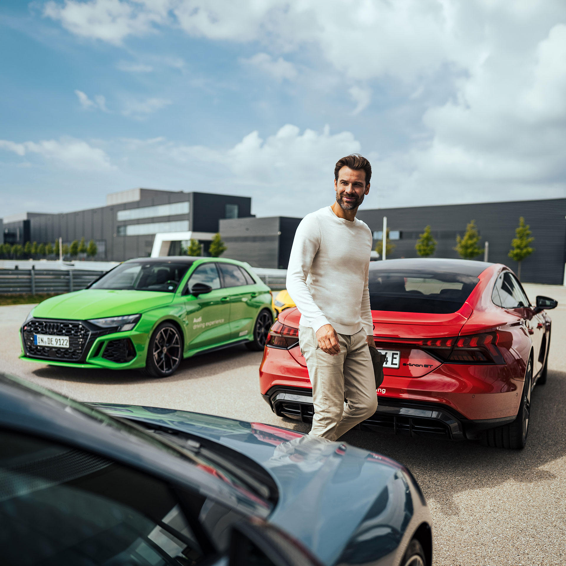 Ein Mann läuft zwischen drei Autos entlang, links steht ein hellgrünes Audi Modell, recht daneben befindet sich ein roter Audi RS e-tron GT und vorne vor befindet sich noch ein weiteres dunkelgraues Audi Modell